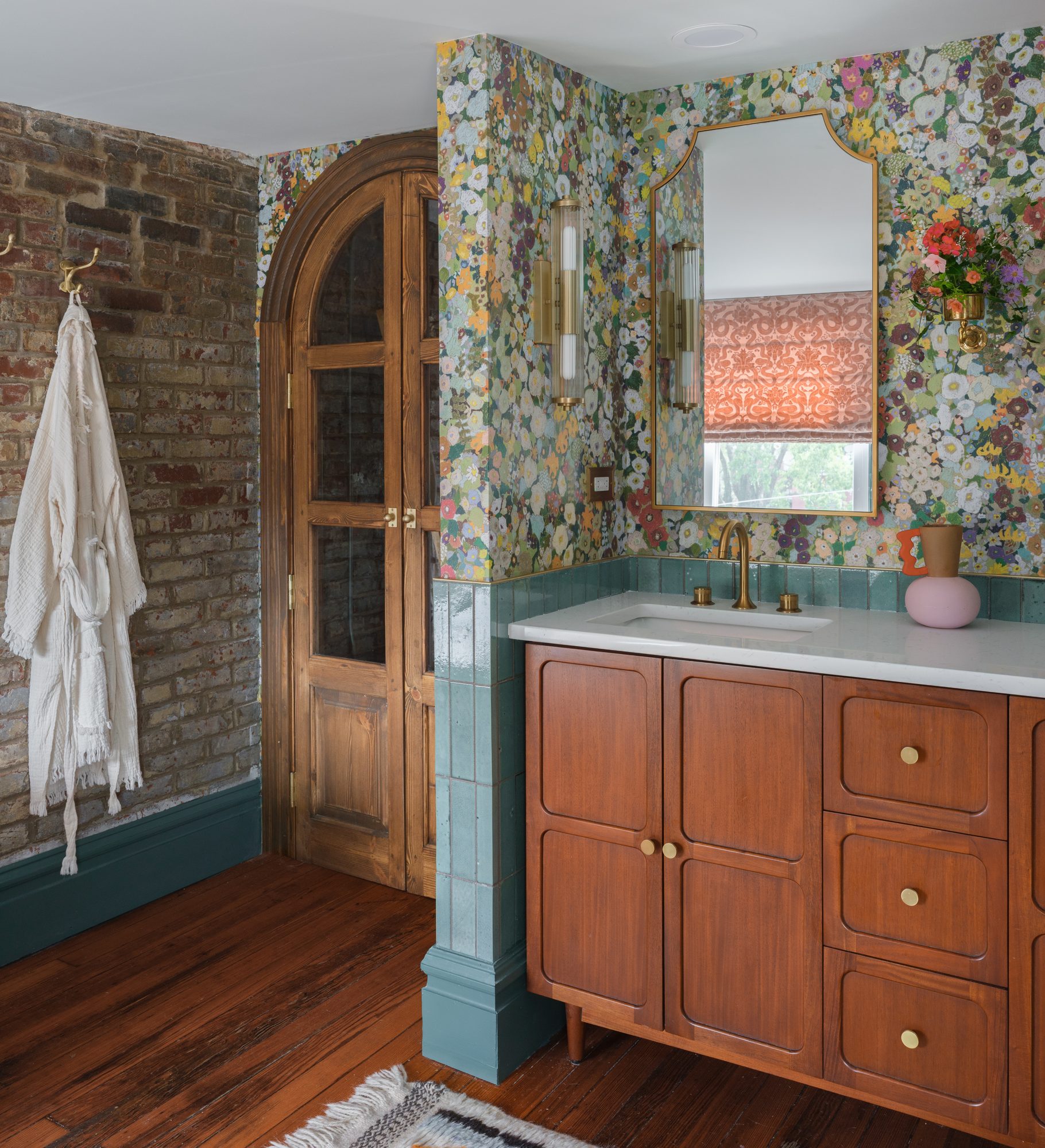 Arched wood doors with glass panels between an exposed brick wall and a wood vanity under floral wallpaper