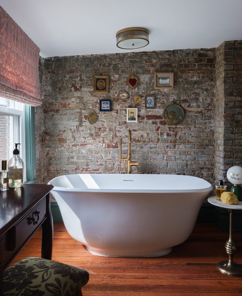 Freestanding tub with brass floor-mounted filler against an exposed brick wall hung with small vintage-style frames in a historic Benton Park bathroom