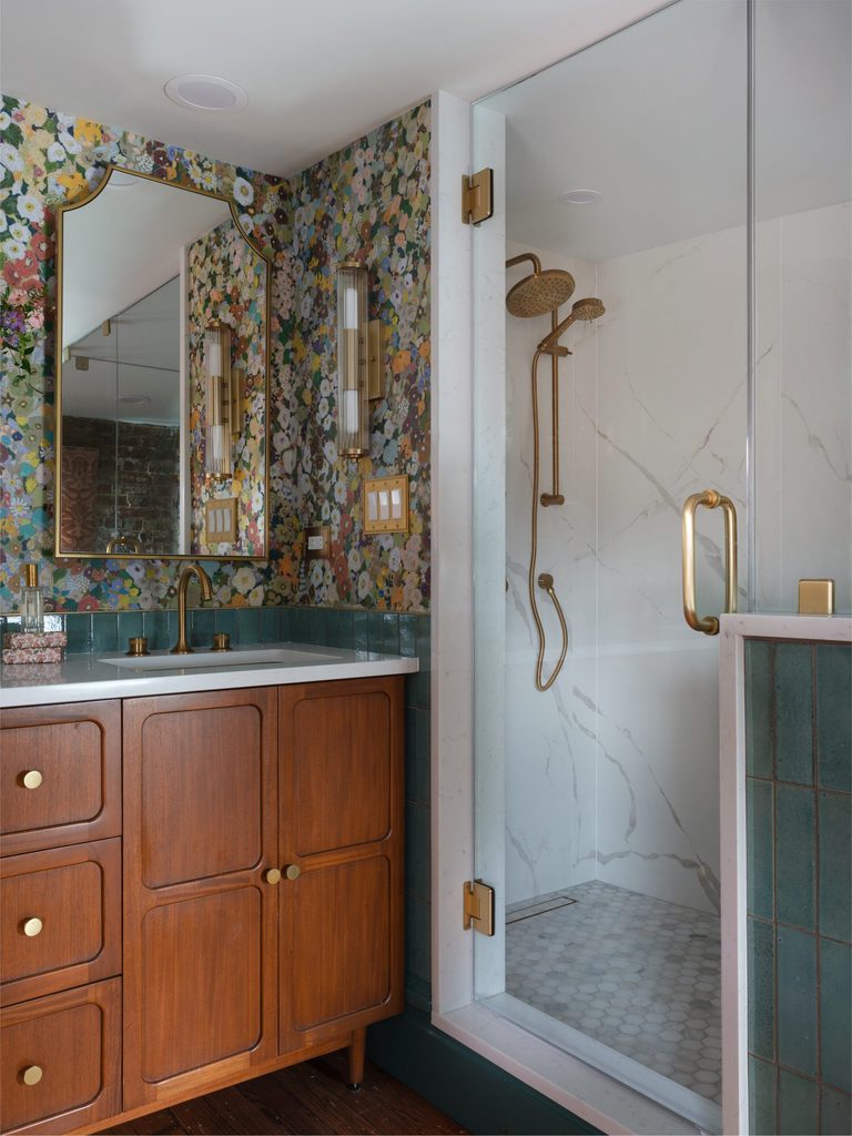 Frameless glass shower with marble-look walls, hex mosaic floor, and brass rain head beside a wood vanity and floral wallpaper