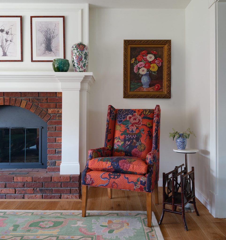 Orange chinoiserie wing chair beside a fireplace under an antique floral painting in a gilt frame