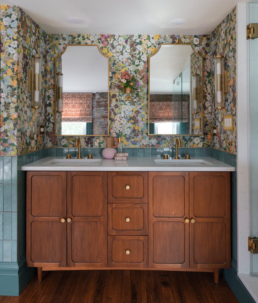 Wood double vanity with rounded panels and brass knobs, arched brass mirrors, fluted sconces, and floral wallpaper over aqua tile